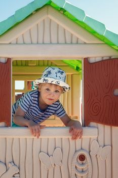A cheerful little boy in a hat, inside a toy little house, he looks out the window with the shutters straight into the camera.