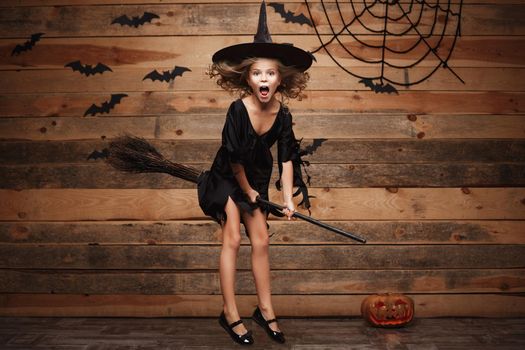 Halloween Witch concept - little caucasian witch child flying on magic broomstick over bat and spider web background.