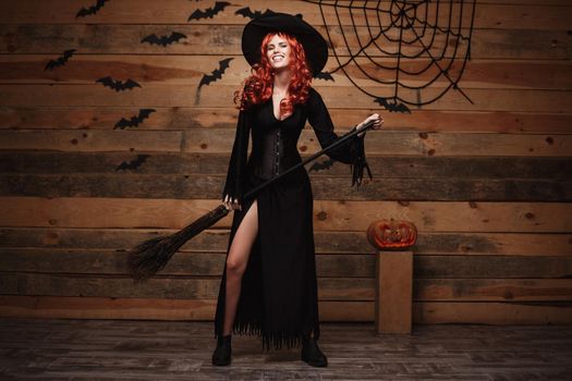 Halloween witch concept - Full-length Happy Halloween red hair Witch holding posing with magic broomstick over old wooden studio background.
