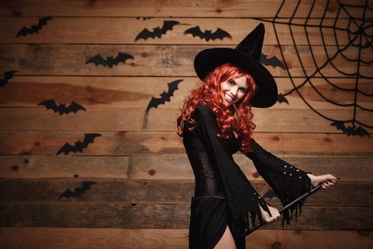Halloween witch concept - Happy Halloween red hair Witch holding magic broomstick flying gesture over old wooden studio background.