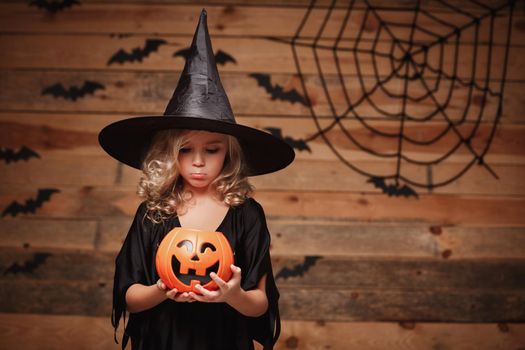 Halloween Witch concept - little caucasian witch child disappointing with no candy in halloween candy pumpkin jar. over bat and spider web background.