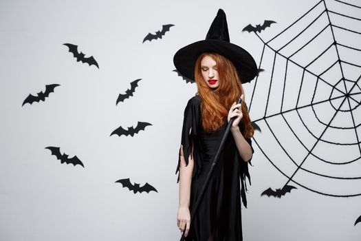 Halloween Witch Concept - Portrait of beautiful young witch with broomstick over grey wall with bat and spider web background.