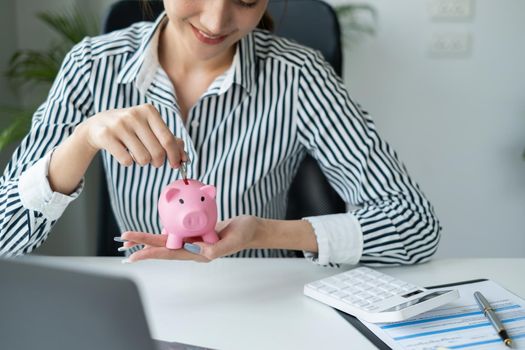 Portrait of Asian business woman smiling putting a coin inside piggy bank as savings for investment. wealth and financial concept.