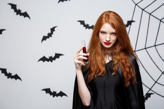 Halloween witch concept - Happy Halloween Witch drinking blood over dark grey studio background with bat and spider web.