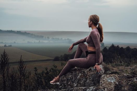 Sporty Young Woman Sitting on the Rock on Top of the Hill Listening Music in Earphones and Looking on Horizon After Sunset