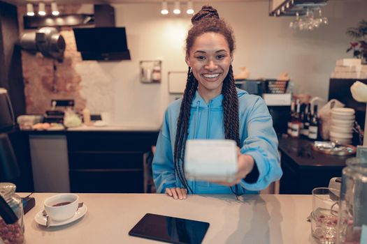 Mixed-races young pretty female barista handing paying terminal to client. Cashless paying.