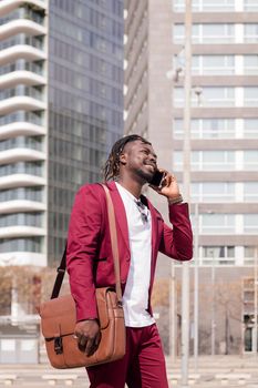 vertical photo of a smiling african businessman with briefcase walks through the city financial downtown talking on phone, concept of technology and communication, copy space for text