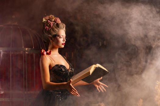Charming woman witch flowers in hair vintage black dress with book of spell in the old castle. Halloween. Celebration.