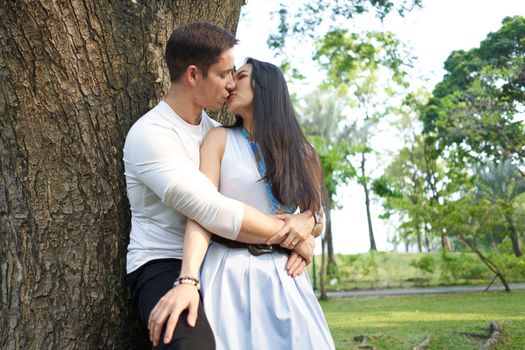 Pofile of a multicultural newlywed couple kissing next to a tree in a park