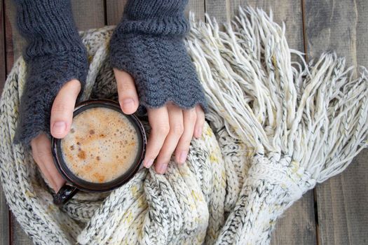 female hands in mittens hold a cup of hot drink with foam. mug of coffee on a wooden table next to a warm knitted scarf. Soft focus.