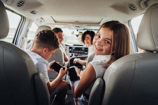 Cheerful Caucasian Teenage Girl Smiles Into the Camera While Sitting in Minivan Car with Her Brother, Mother and Father, Happy Four Members Family Enjoying Weekend Road Trip