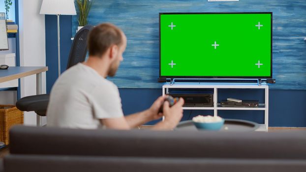 Man holding wireless controller playing console video game on green screen tv while sitting on sofa
