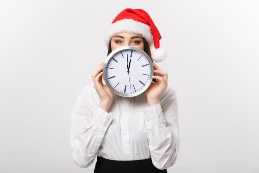 Time management concept - Young business woman with santa hat hiding behind a clock isolated over white background.