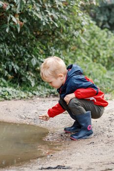 A child in a red jacket and rubber boots explores the mud of a swamp in nature on a hike in the fresh air