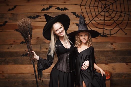 Halloween Concept - cheerful mother and her daughter in witch costumes celebrating Halloween posing with curved pumpkins over bats and spider web on Wooden studio background.
