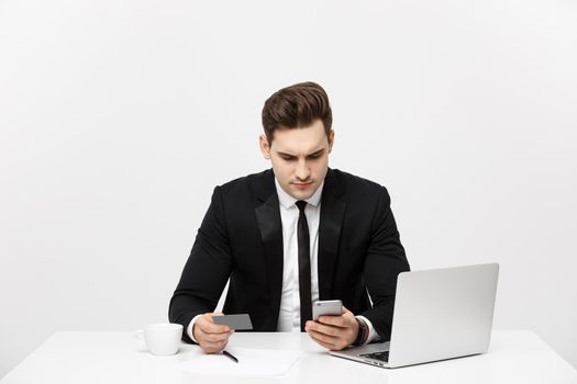 Business Concept: Portrait of young businessman using laptop computer and mobile phone holding debit card. Isolated over grey background.