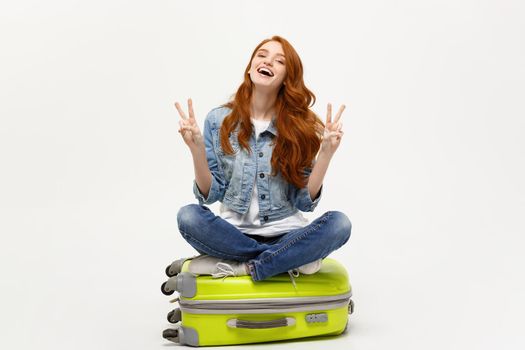 Travel concept: young smiling caucasian woman siting on suitcase showing two fingers. Isolated over white background.