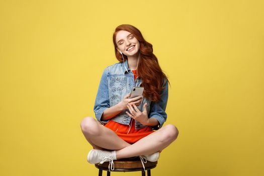 Lifestyle Concept: Pretty girl with long curly red hair enjoy listening to music on her phone and sitting on wooden chair on vivid yellow background in studio.