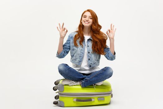 Travel and Lifestyle Concept: Young caucasian Woman sitting on suitcase and showing ok finger sign. Isolated on white.