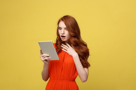 Technology and Lifestyle Concept: Surprised young woman wearing orange dress clothes using tablet pc isolated on vivid yellow background
