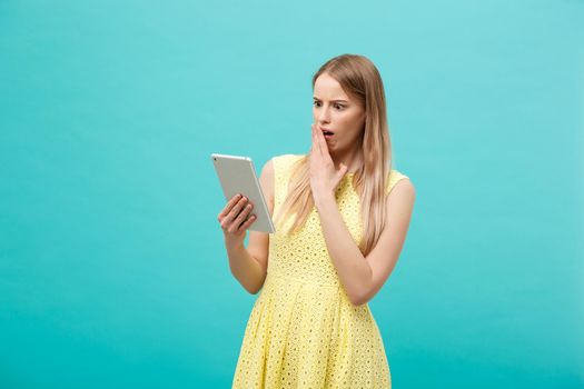 Portrait of a surprised amazed young woman looking at digital tablet isolated on a blue studio background.