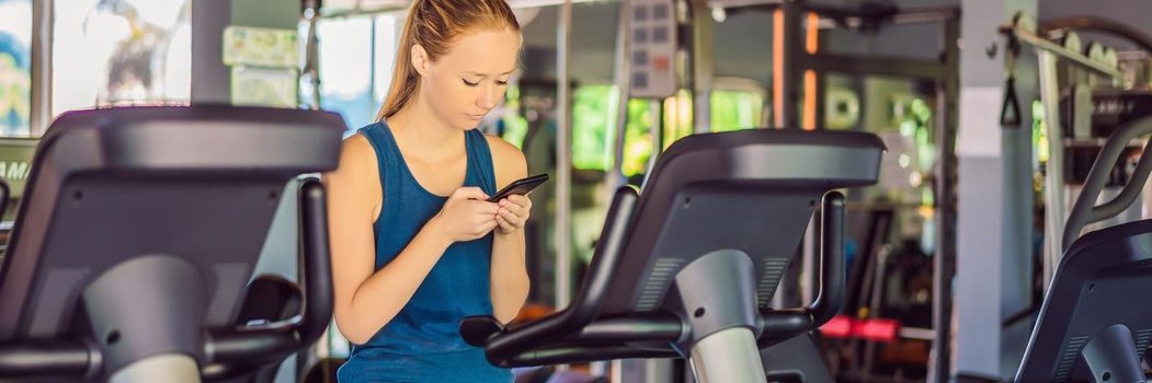 Young woman using phone while training at the gym. Woman sitting on exercising machine holding mobile phone BANNER, LONG FORMAT