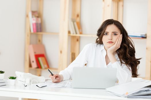 Tired beautiful Businesswoman holding hand on head while working on computer and some business documents in bright office