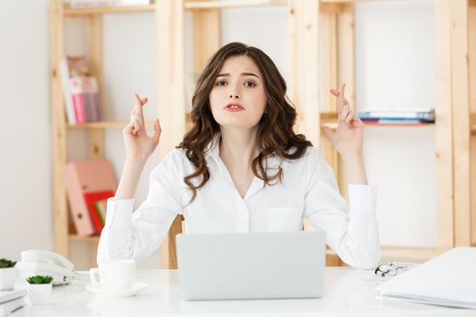 Portrait of happy young caucasian business woman sitting at the office desk and holding fingers crossed isolated over white background