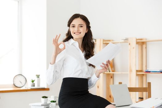 Businesswoman in formal wear holding documents and showing ok sign at modern office, copy space.