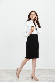 Full length Portrait of beautiful young business woman in formal wear walking and talking for cell phone with takeaway coffee in hand isolated over gray background