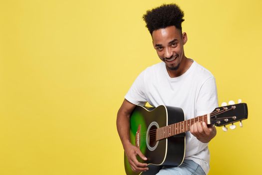 Young handsome African American retro styled guitarist playing acoustic guitar isolated on yellow gold background.