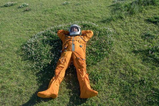 Happy cosmonaut wearing space suit and helmet having a rest while lying on green grass outdoor