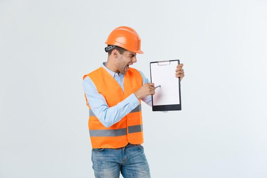 Frustrated young engineer with hardhat and reflective vest checking on mistake in document over gray background
