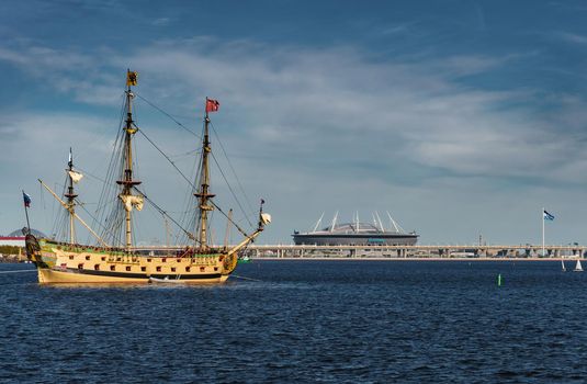 Russia, St. Petersburg, 20 May 2022: An ancient sailing frigate sails on the calm sea in sunset, the new stadium Gazprom arena by the oil company Gazprom on background