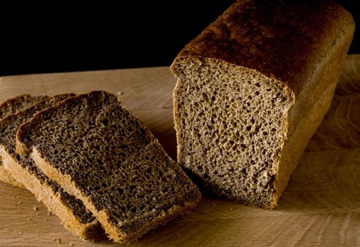 Loaf of black rye bread, cut into pieces, in the shape of a brick on a wooden board on a black background