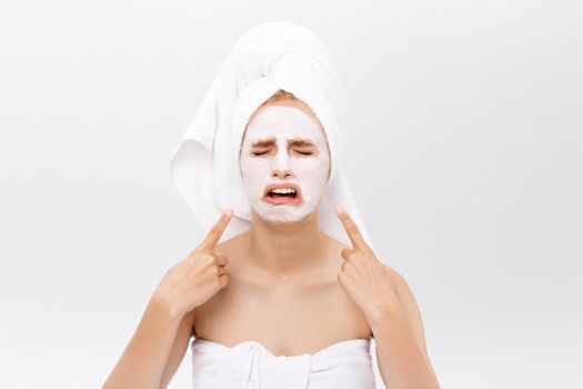 Unhappy beautiful young woman with white clay face mask makes acne therapy, wears white towel on hair, posing against white studio wall. Horizontal shot of Caucasian female with organic facial mask