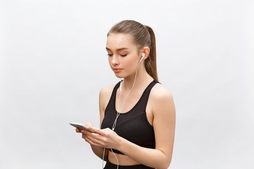 Isolated shot of young sportswoman has healthy athleteic body, listens music with headphones, holds smart phone. Isolated over grey background.