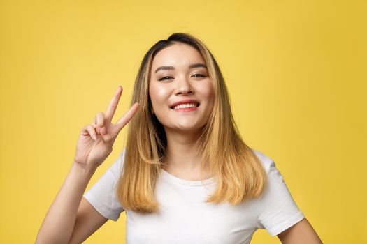Pleased asian woman in t-shirt showing peace gestures and looking at the camera over yellow background