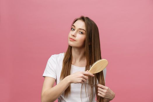 Smiling young woman combing hair and looking away isolated on pink