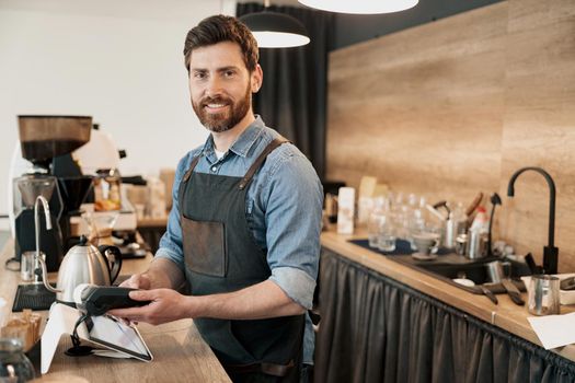 Smiling barista holds a payment terminal in his hands and looks at the camera