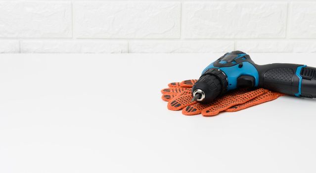 a pair of orange textile work gloves and a portable drill on a battery lie on a white table against a background of a white brick wall
