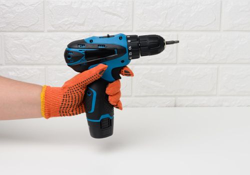 hand in a textile glove holds a portable drill on a battery against a background of a white brick wall