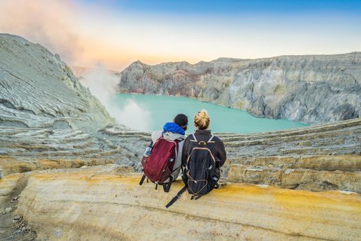 Young tourist man and woman sit at the edge of the crater of the Ijen volcano or Kawah Ijen on the Indonesian language. Famous volcano containing the biggest in the world acid lake and sulfur mining spot at the place where volcanic gasses come from the volcano. They enjoy the magnificent view on the crater