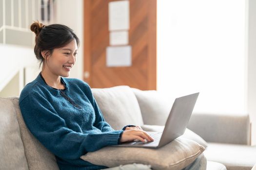 Smiling young asian woman using laptop, sitting on couch at home