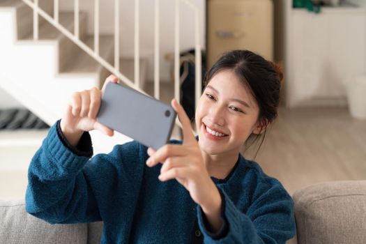 Photo of young asian woman taking selfie on cell phone while sitting on couch in bright apartment.