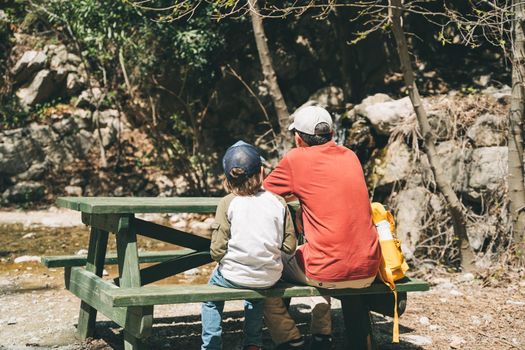 Rear view of tourists school boy and his dad sitting at the wooden table when taking a hike. Child boy and father wearing casual clothes and yellow backpack taking rest on a mountain hike