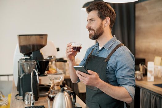 barista wearing apron drinking coffee and holding phone during lunch break