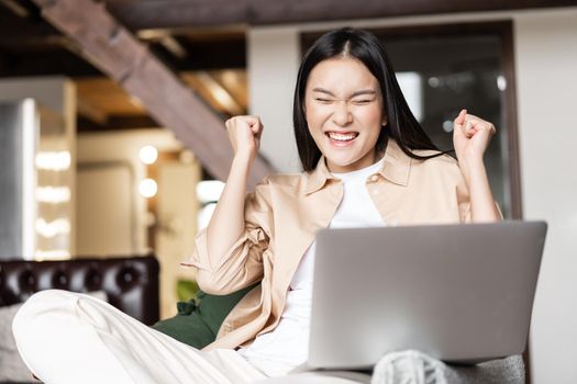 Asian woman celebrating, cheering from good news on laptop, triumphing from joy, sitting in living room at home