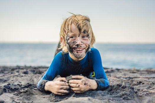 Black Friday concept. Smiling boy with dirty Black face sitting and playing on black sand sea beach before swimming in ocean. Family active lifestyle, and water leisure on summer vacation with kids. Black Friday, sales of tours and airline tickets or goods