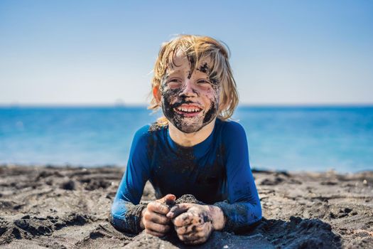 Black Friday concept. Smiling boy with dirty Black face sitting and playing on black sand sea beach before swimming in ocean. Family active lifestyle, and water leisure on summer vacation with kids. Black Friday, sales of tours and airline tickets or goods